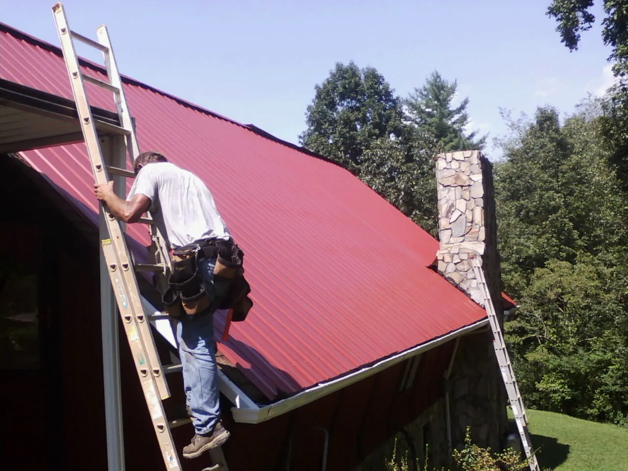 Roofer on a ladder beside a red metal roof and stone chimney — Soffit & Fascia Repair by Roofing Brothers in Shawnee